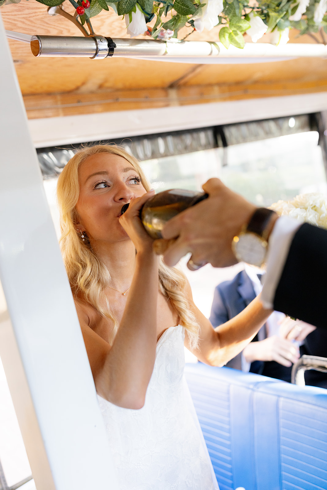 Bride sips champagne straight from the bottle, handed by the groom, celebrating their fun-filled trolley ride on their wedding day.