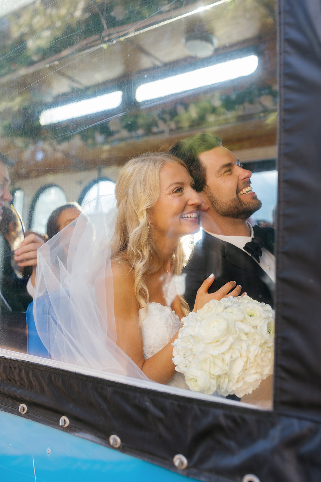 Bride and groom laugh together on a trolley, leaning against the window as golden sunlight reflects their happiness.