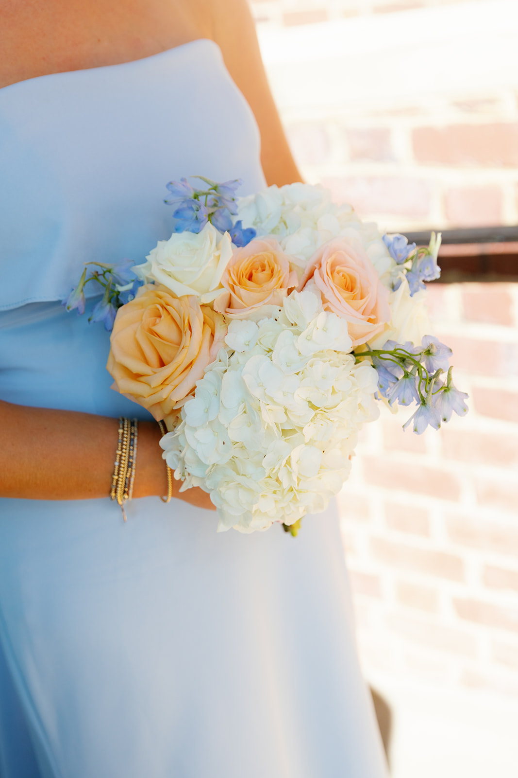 Bridesmaids in elegant light blue dresses hold lush bouquets of peach and white roses.