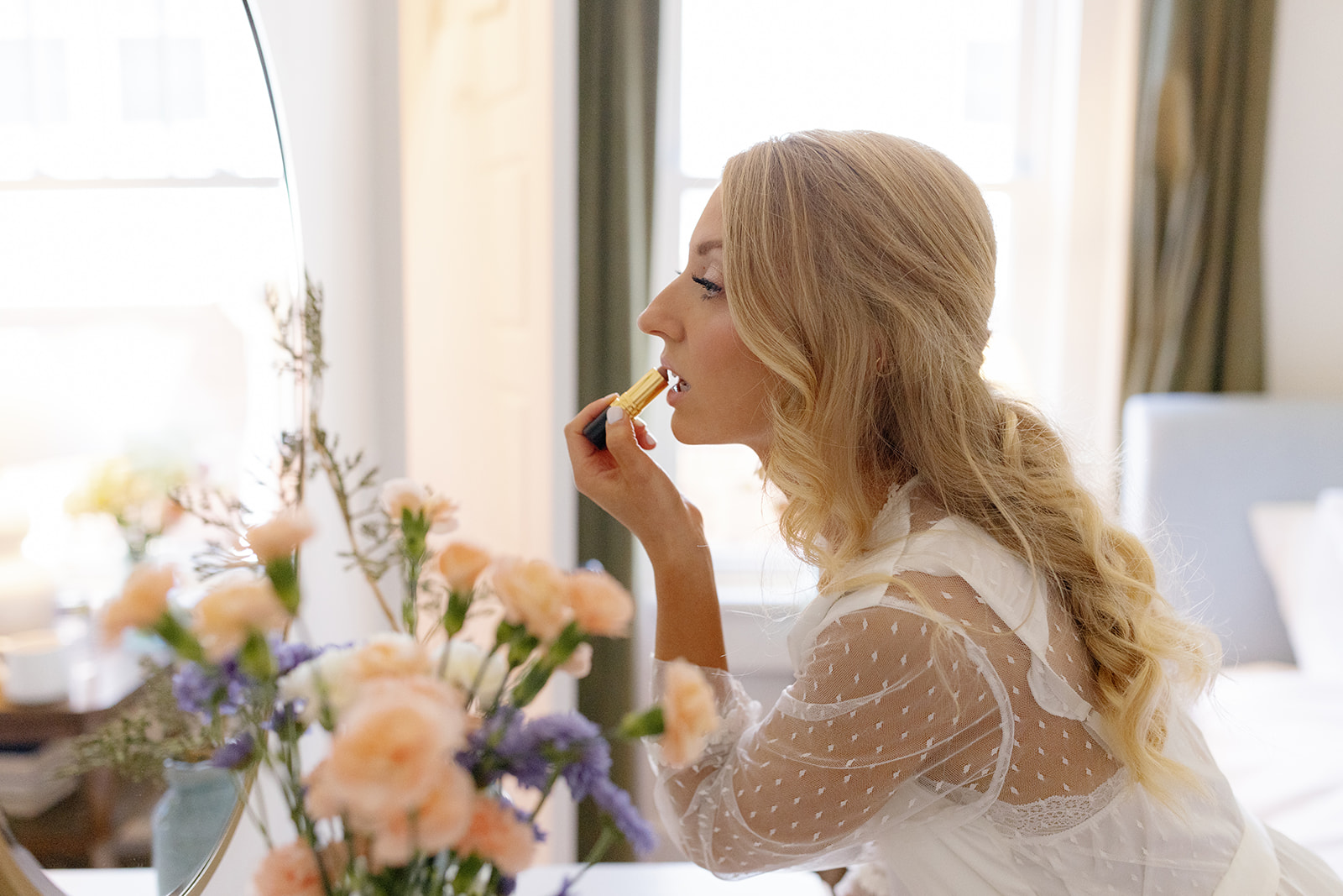 Bride applying lipstick in front of a softly lit mirror, surrounded by delicate florals.
