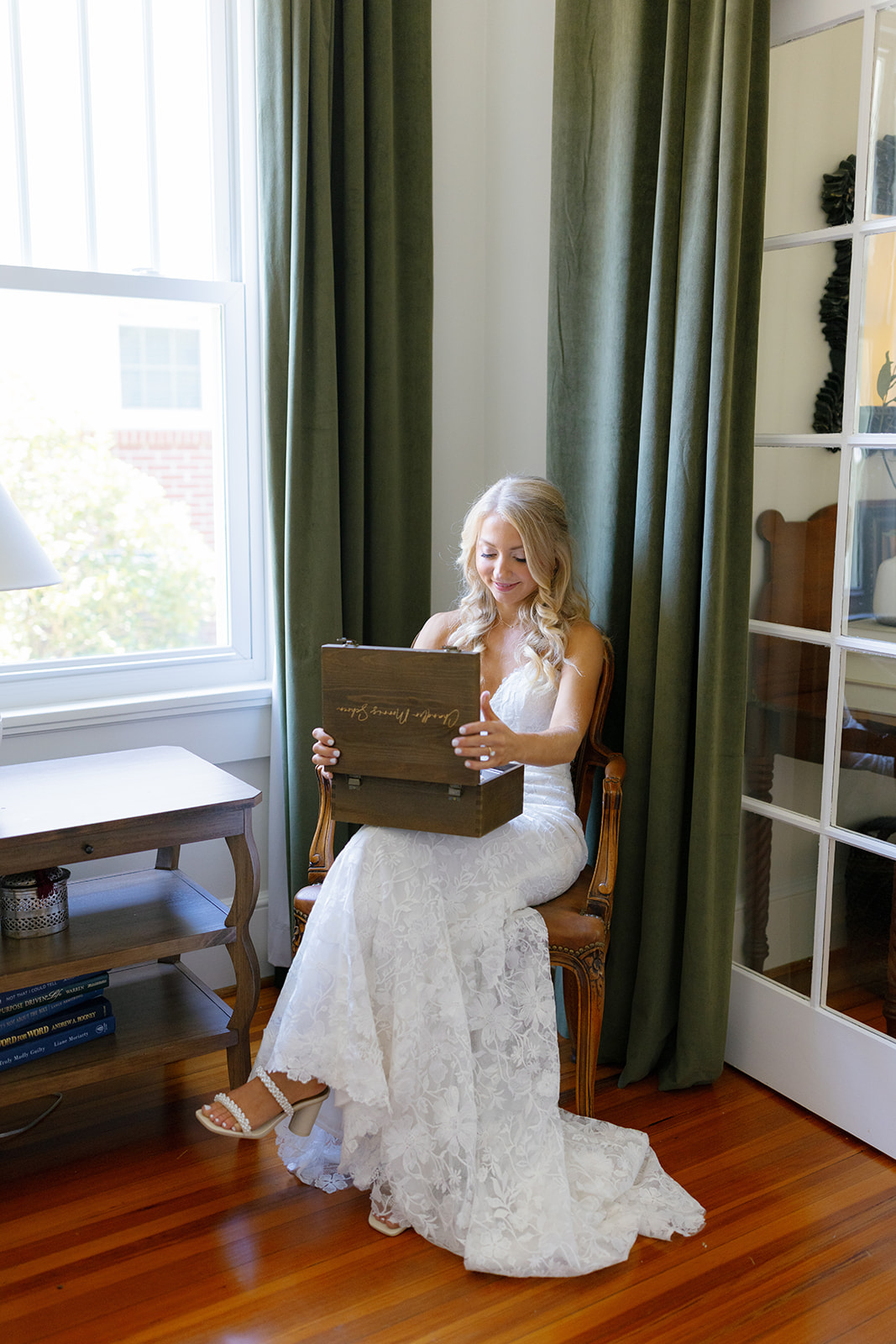 Bride in lace wedding gown sitting by a window, opening a heartfelt gift from her groom.