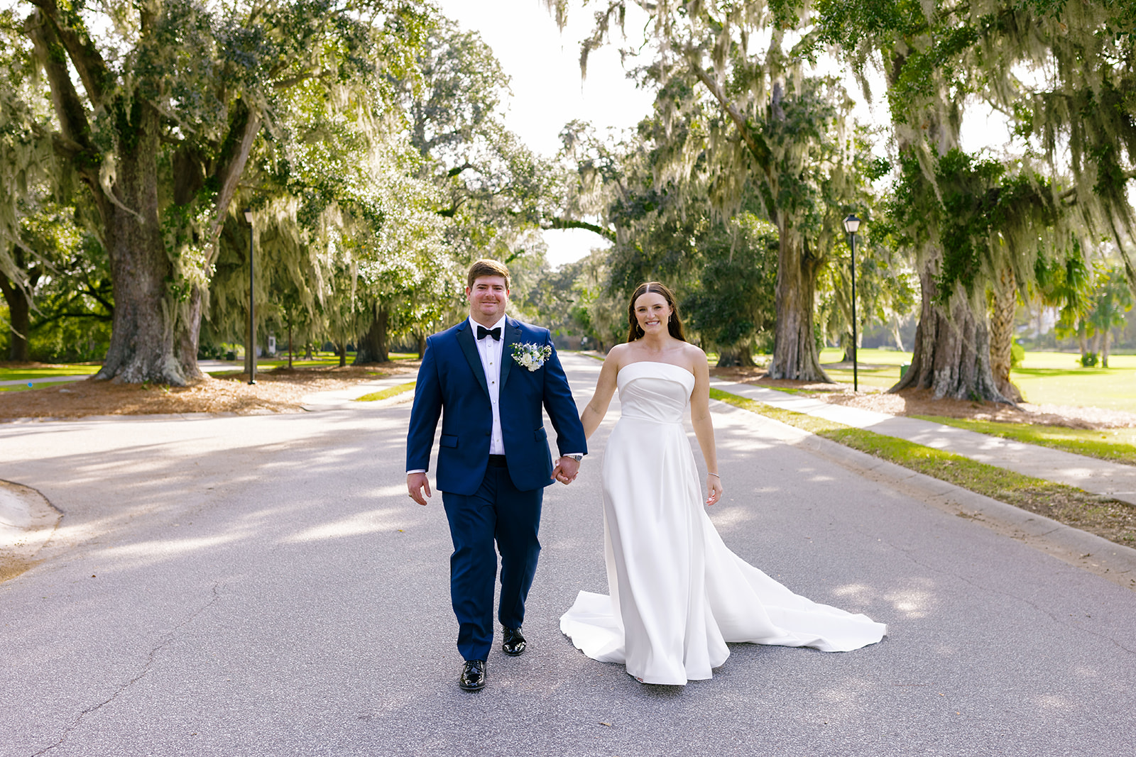 couple walk hand in hand beneath Charleston oaks in their wedding attire.