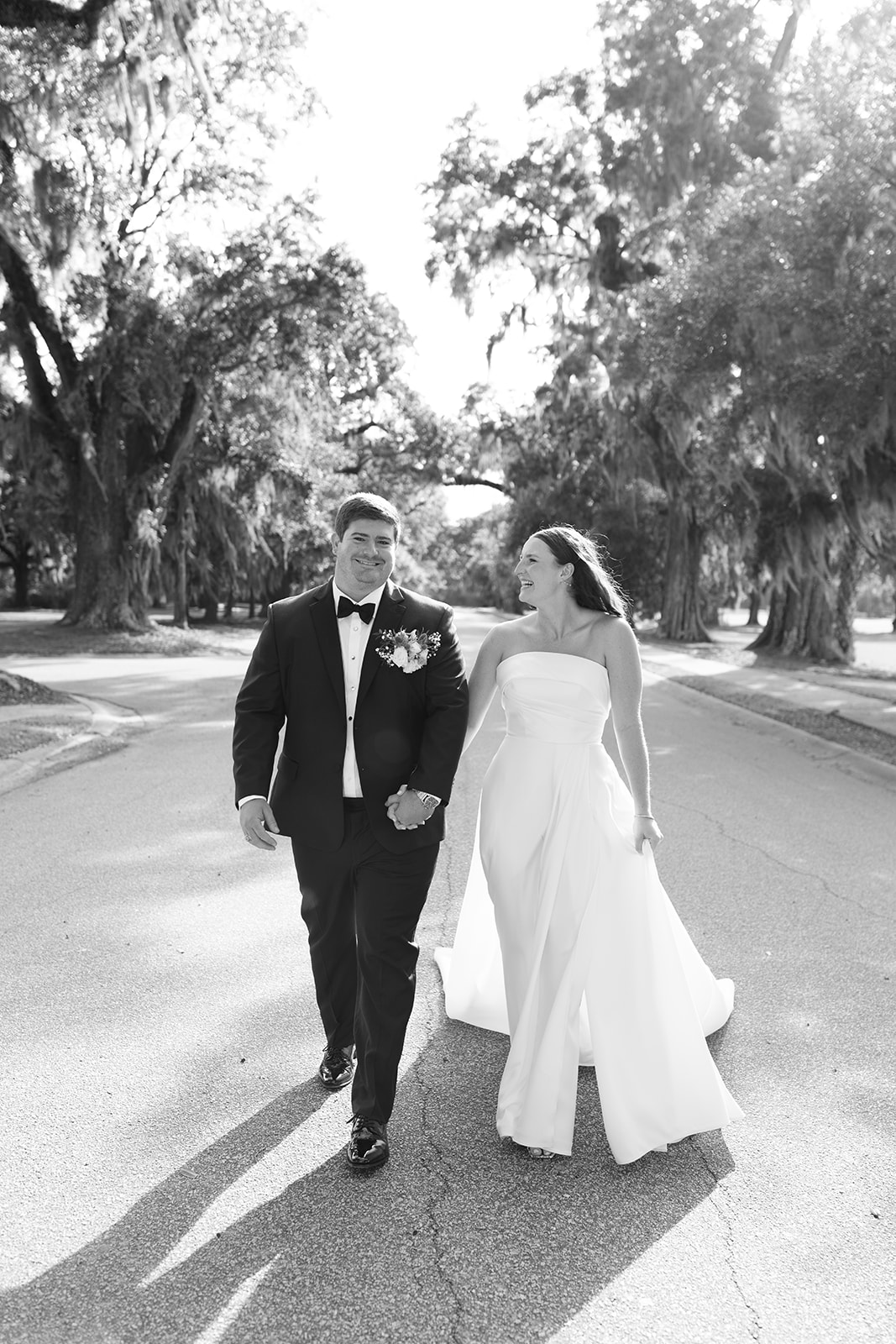 The couple walks hand in hand, laughing together beneath the trees at Dunes West.