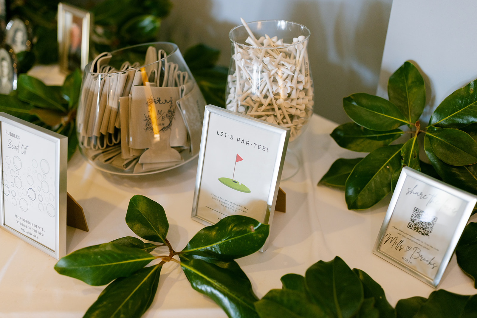 Wedding favor table decorated with golf tees, custom koozies, and signs, styled with magnolia leaves.