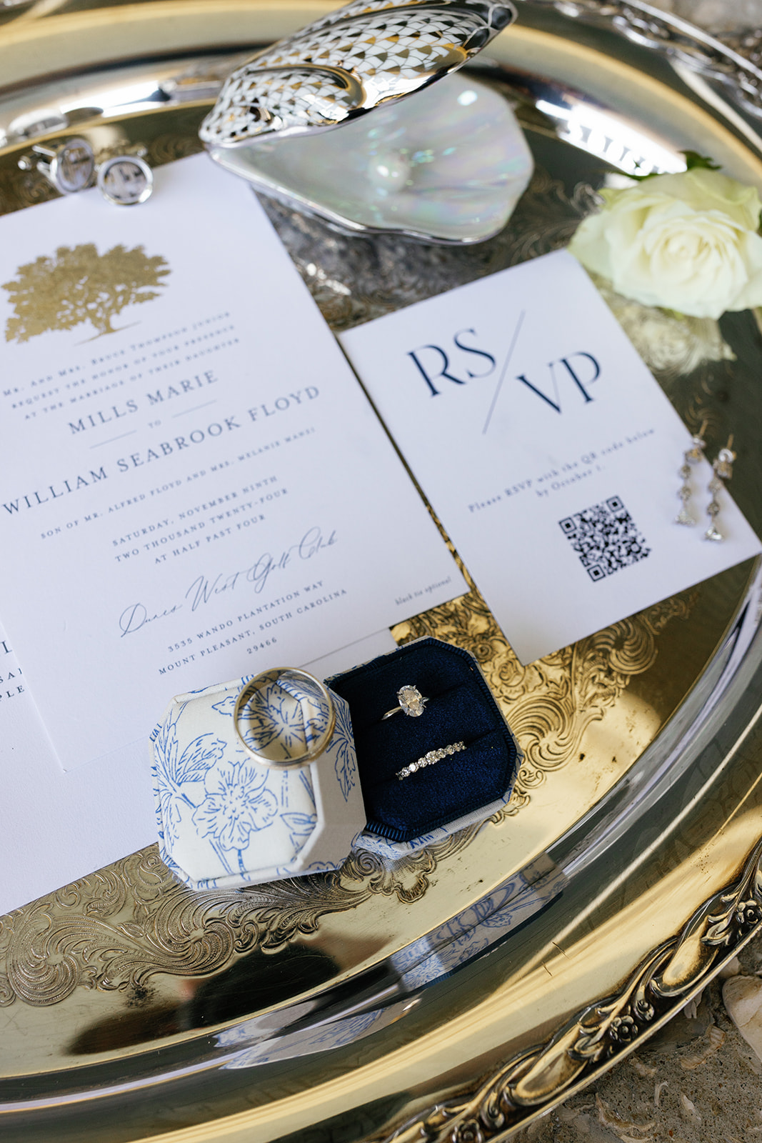 Flat lay of wedding invitation suite, RSVP card, rings, and bridal jewelry on a silver tray with white rose accents.
