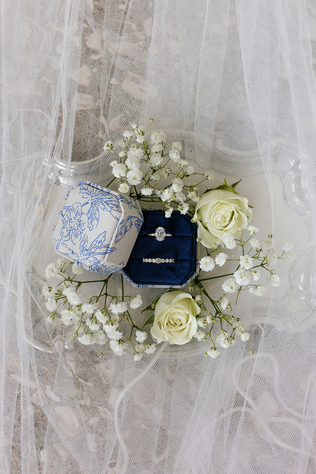 Bridal rings displayed in blue velvet and floral boxes, surrounded by white roses and baby’s breath under the veil.