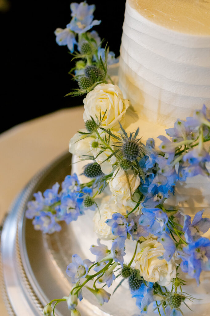 Close-up of a white wedding cake adorned with delicate blue delphinium, white roses, and thistle accents.