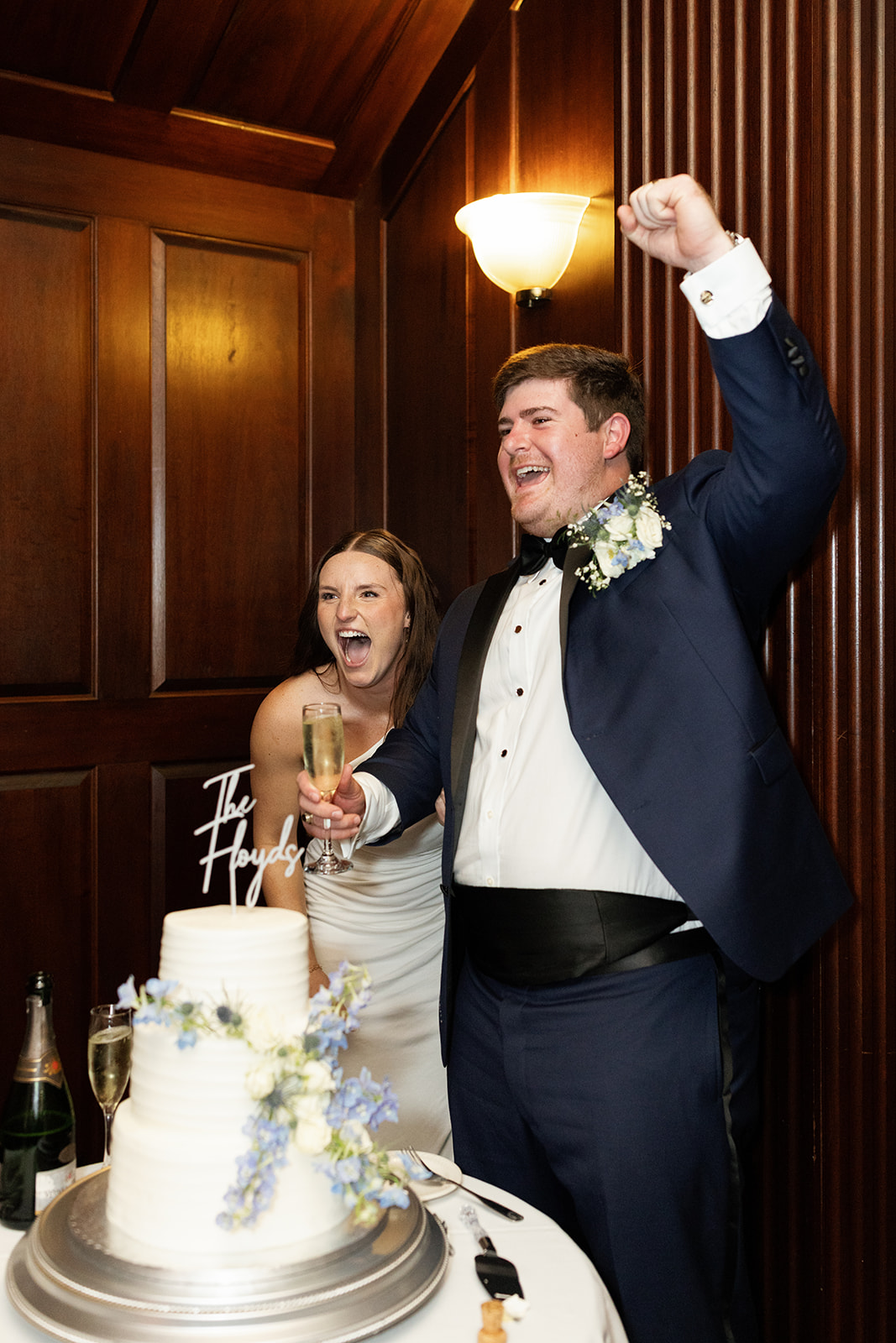 Couple celebrates joyfully with raised arms and bubbly next to their three-tier wedding cake.