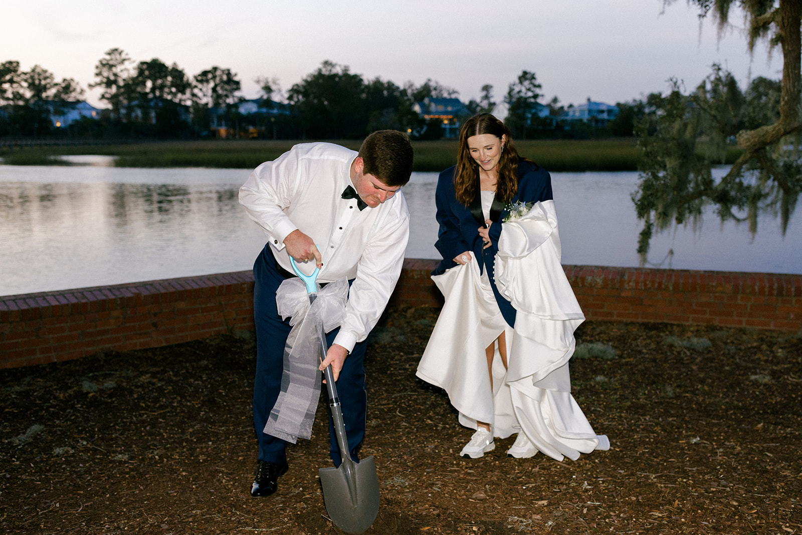 Groom unearths the buried bourbon bottle while the bride watches and laughs near the water’s edge.