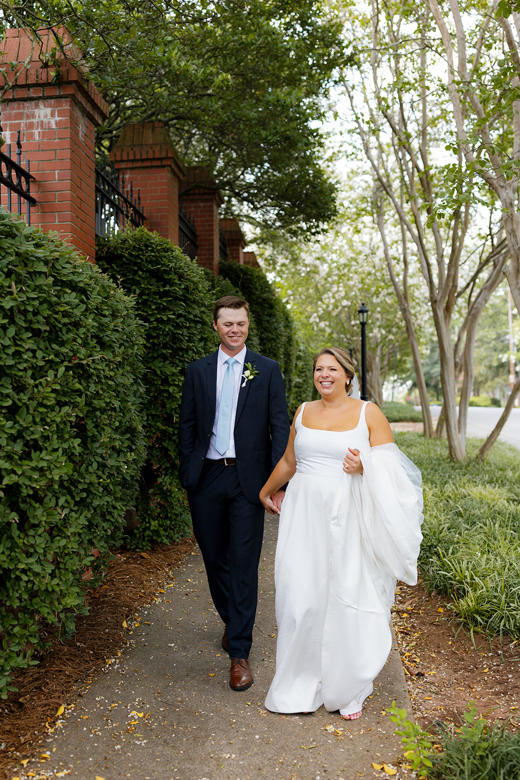 The couple walks hand in hand along a garden path, smiling and relaxed under the trees.