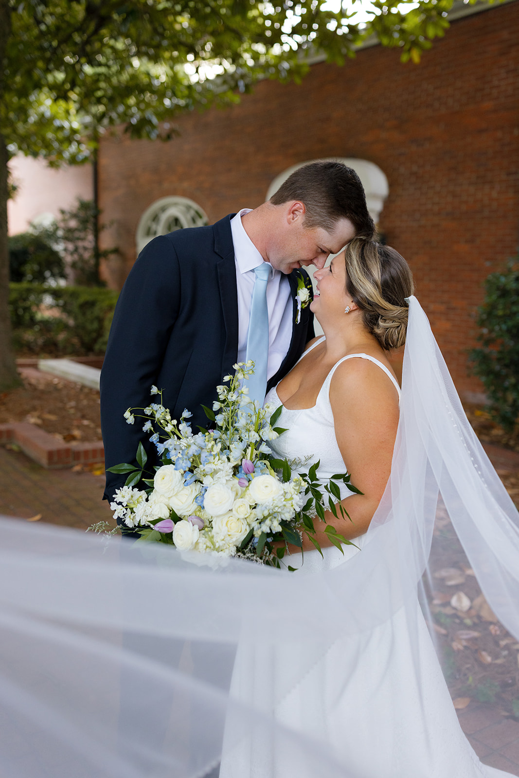 Newlyweds share a quiet, romantic moment, foreheads touching as her veil flows around them.