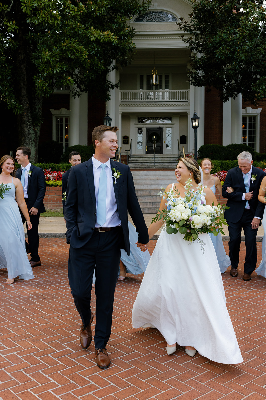 The couple walks hand in hand, smiling with their wedding party on the red brick path in front of the Poinsett Club.