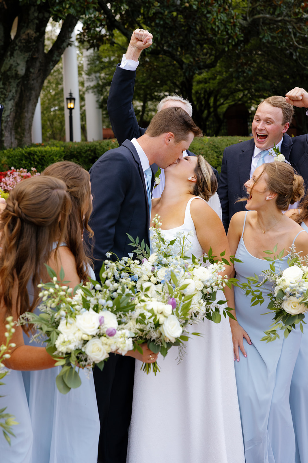 The pair shares a joyful kiss surrounded by cheering bridesmaids and groomsmen holding lush bouquets.