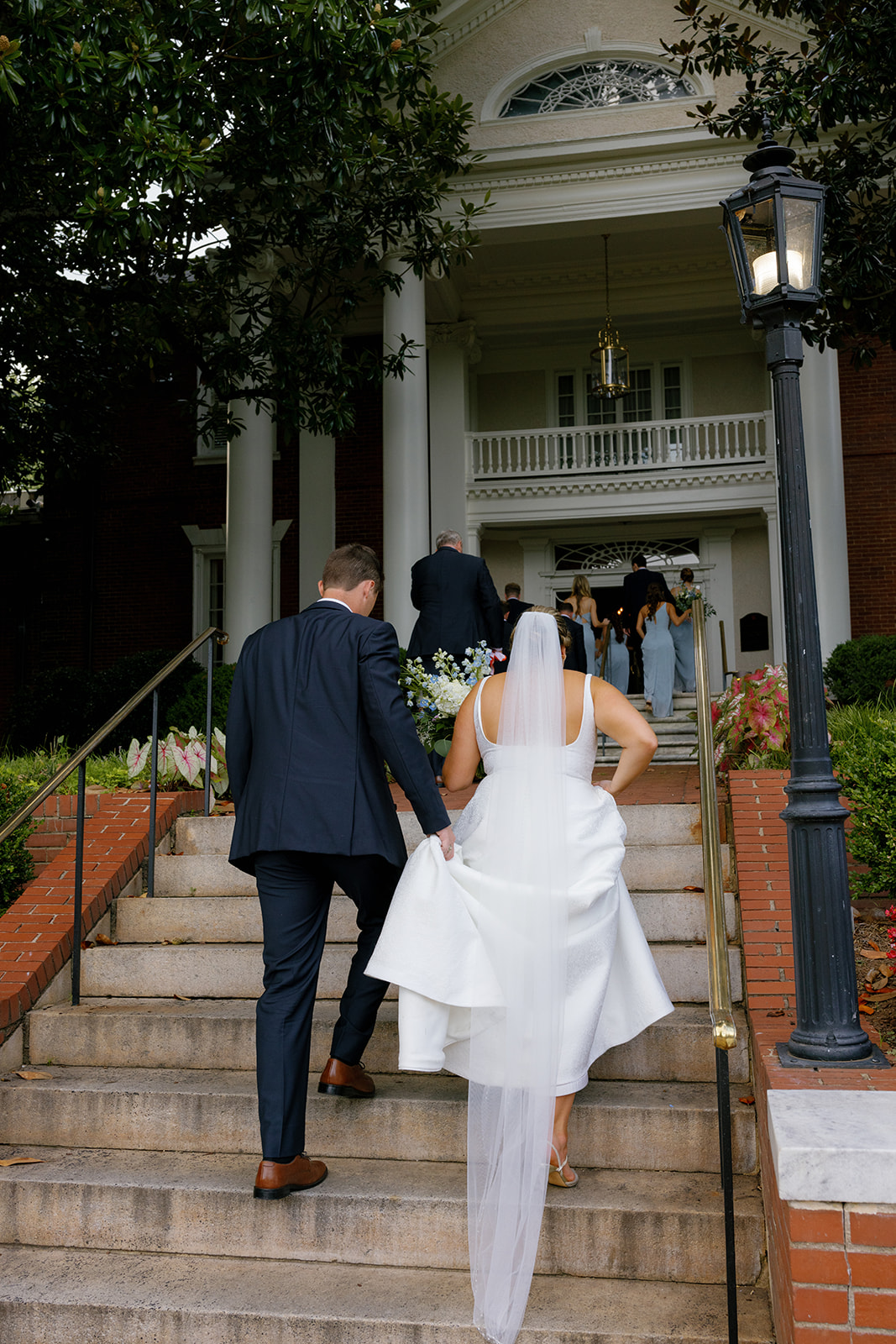 Bride and groom walk up the grand staircase, the bride lifting her dress slightly with bouquet in hand.