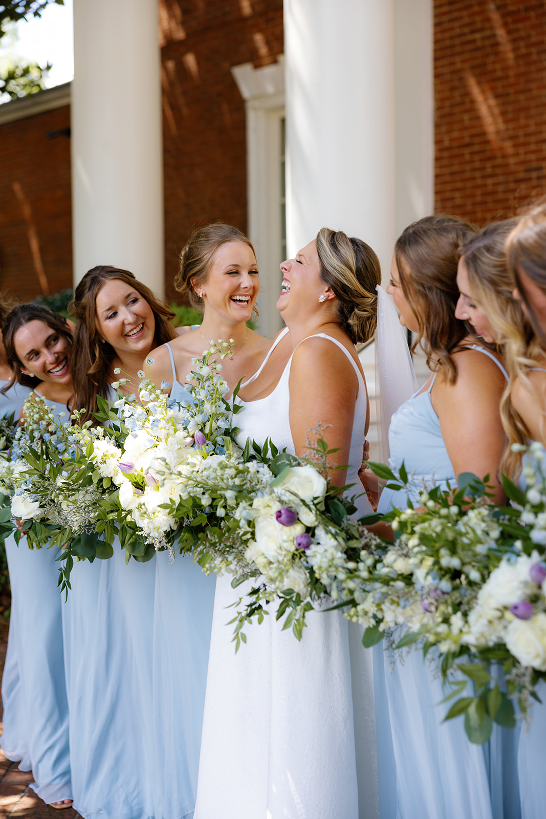 Bride laughs with her bridal party dressed in dusty blue, bouquets full of white and purple blooms taken by a Greenville wedding photographer.