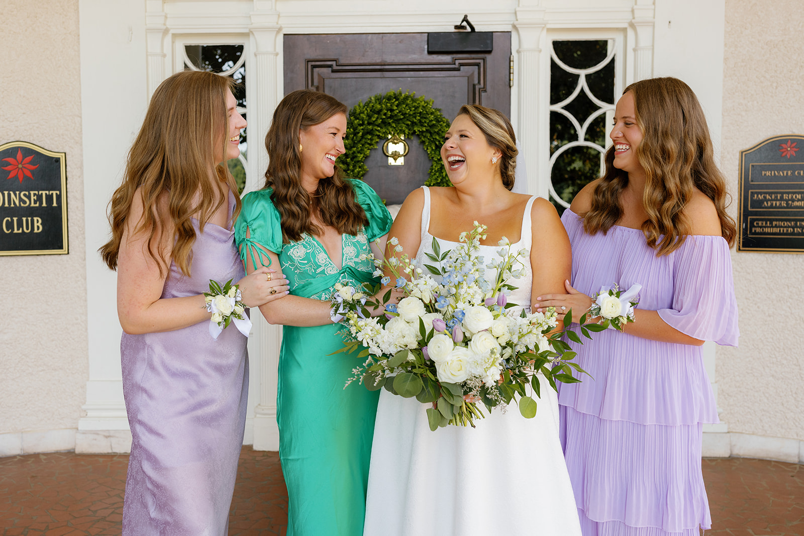 Bride and bridesmaids laugh together while holding lush spring bouquets outside the venue doors.