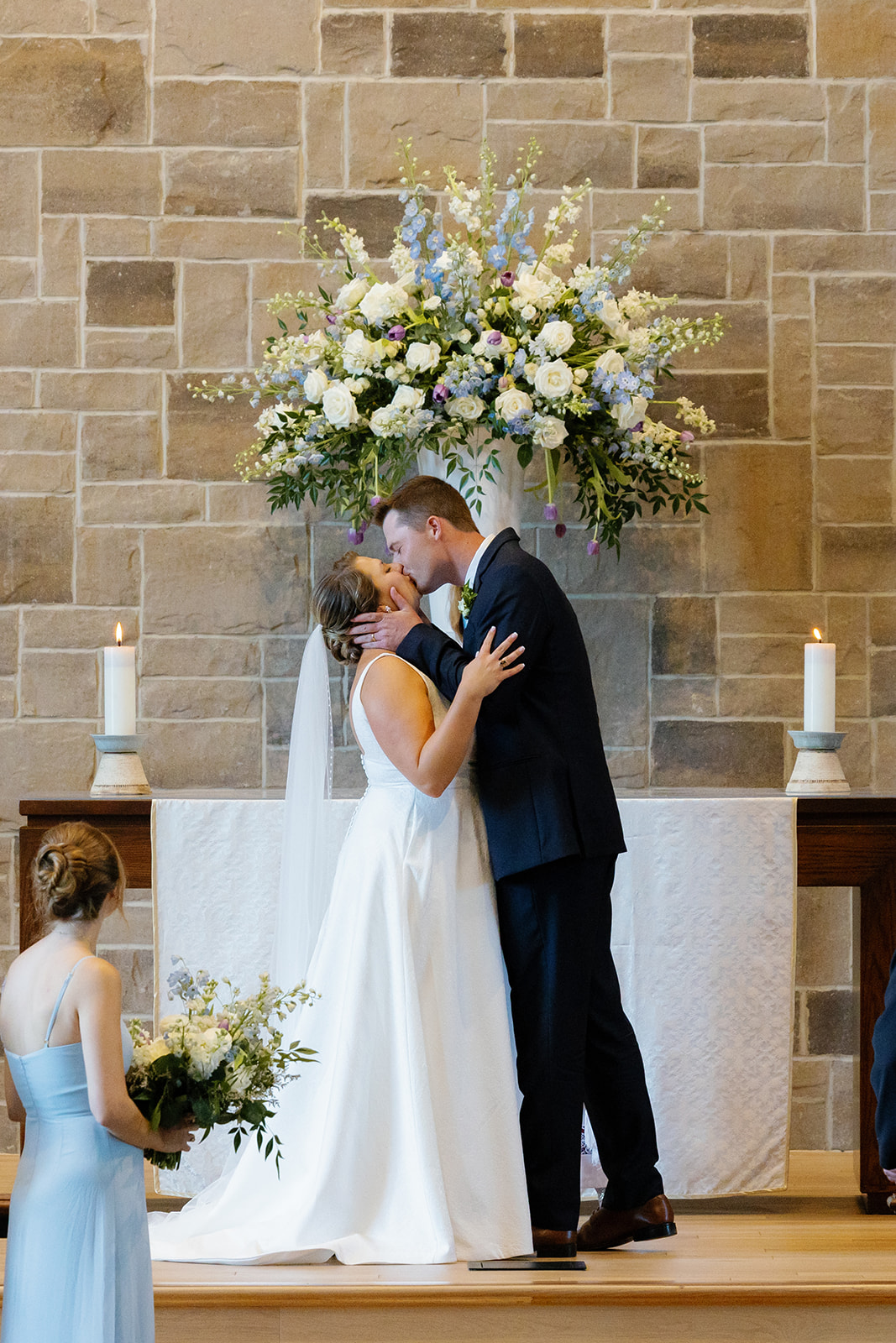 Newlyweds share a romantic first kiss in front of a towering floral display of white and lavender blooms.