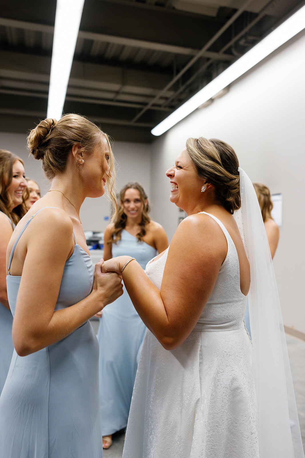 Bride laughs joyfully while holding hands with a bridesmaid, surrounded by the bridal party in soft blue gowns.
