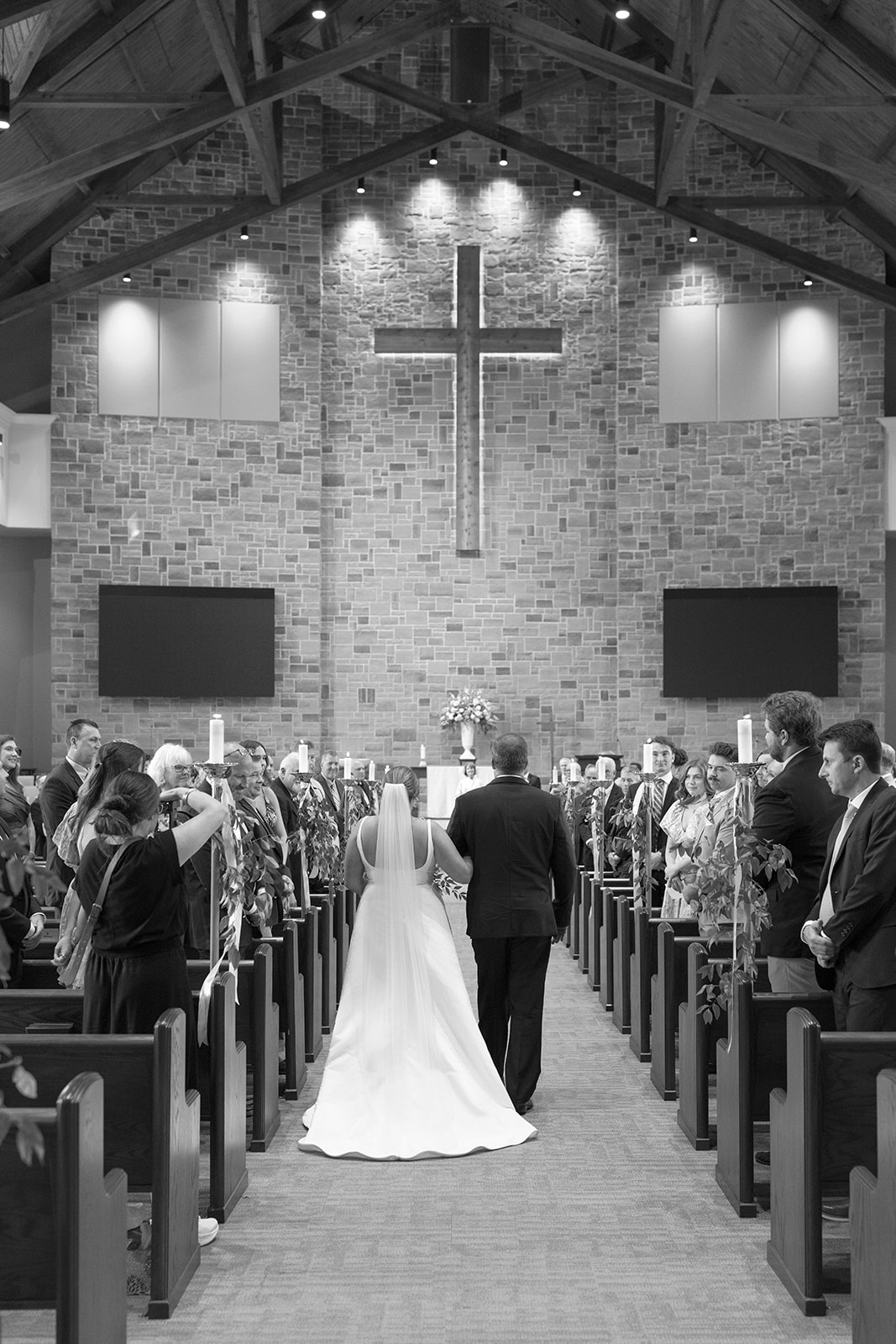 Bride and her father walk down the aisle toward the altar under vaulted wooden beams and soft candlelight.