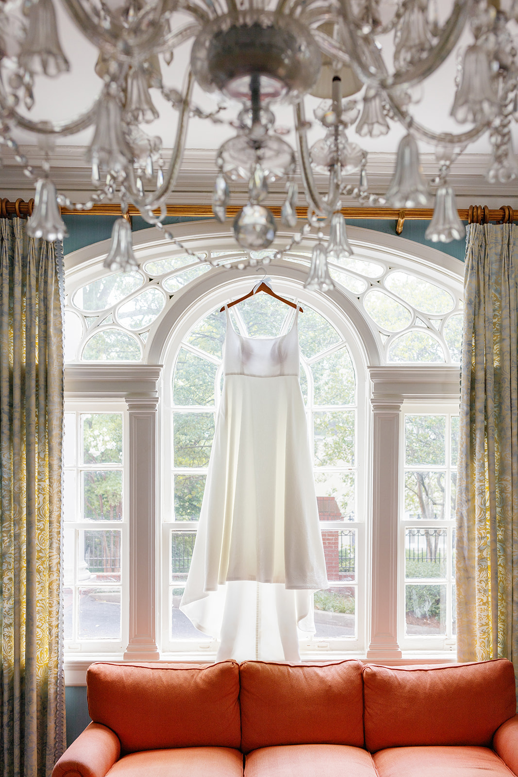 bride’s classic satin wedding gown hangs in a sunlit window framed by vintage chandeliers and arched glass.