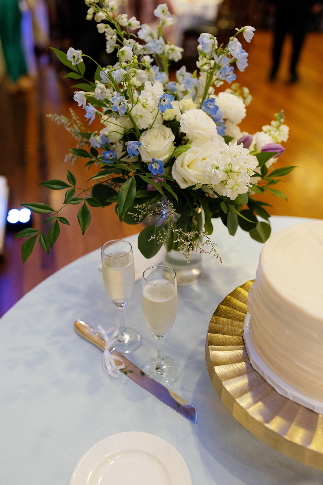 Romantic cake table with champagne flutes, ivory tiered cake, and soft blue and white floral arrangement.