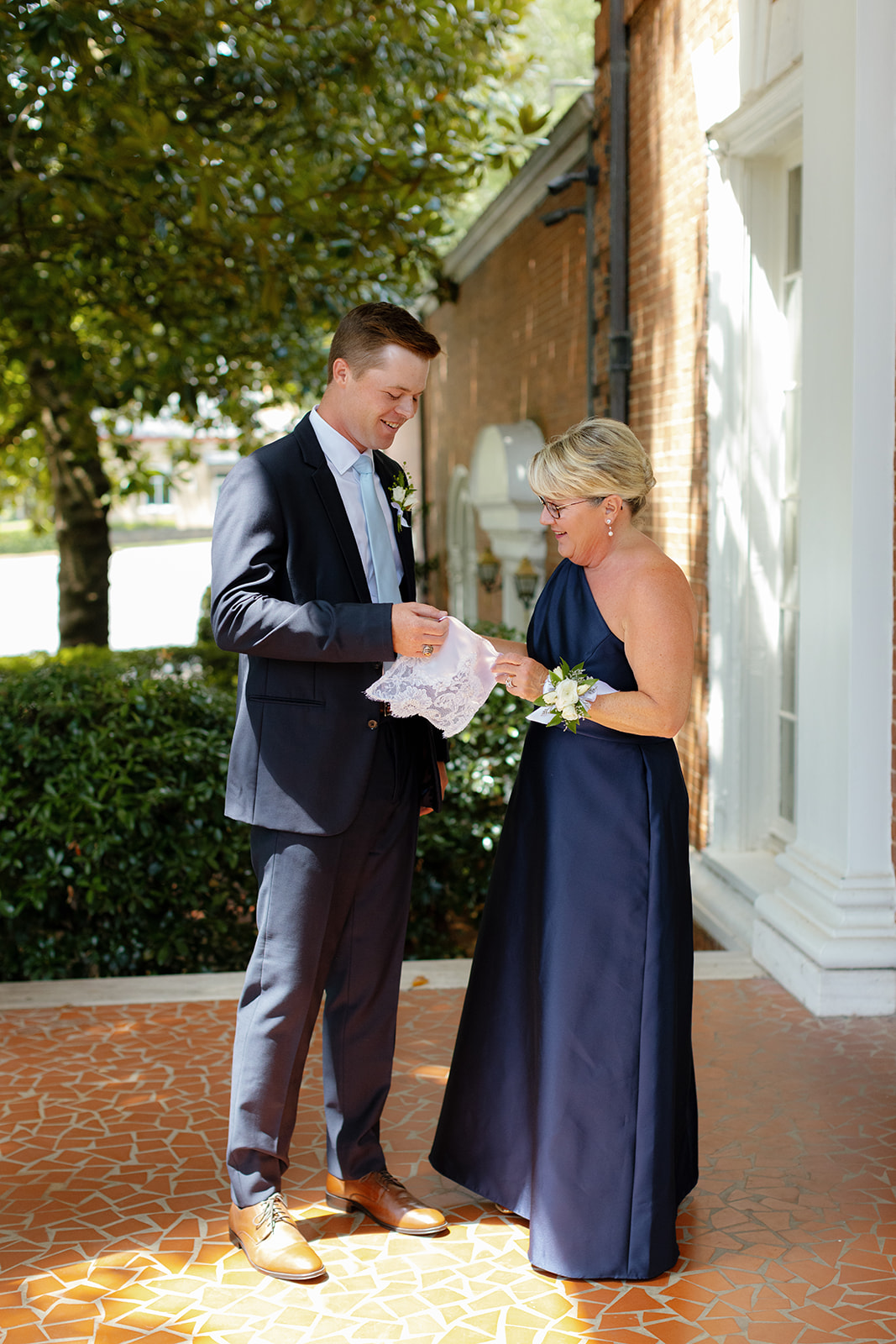 Groom and his mother smile warmly while exchanging a lace handkerchief during a quiet moment together taken by a Greenville wedding photographer.