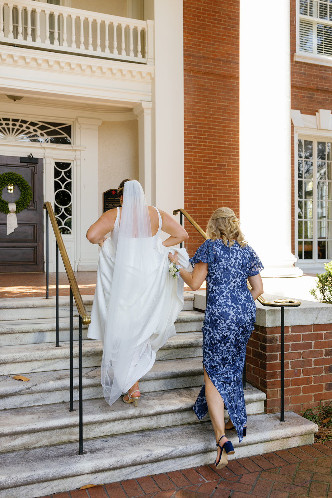 Bride walks up marble steps with her mother, holding her gown as they enter the ceremony site.