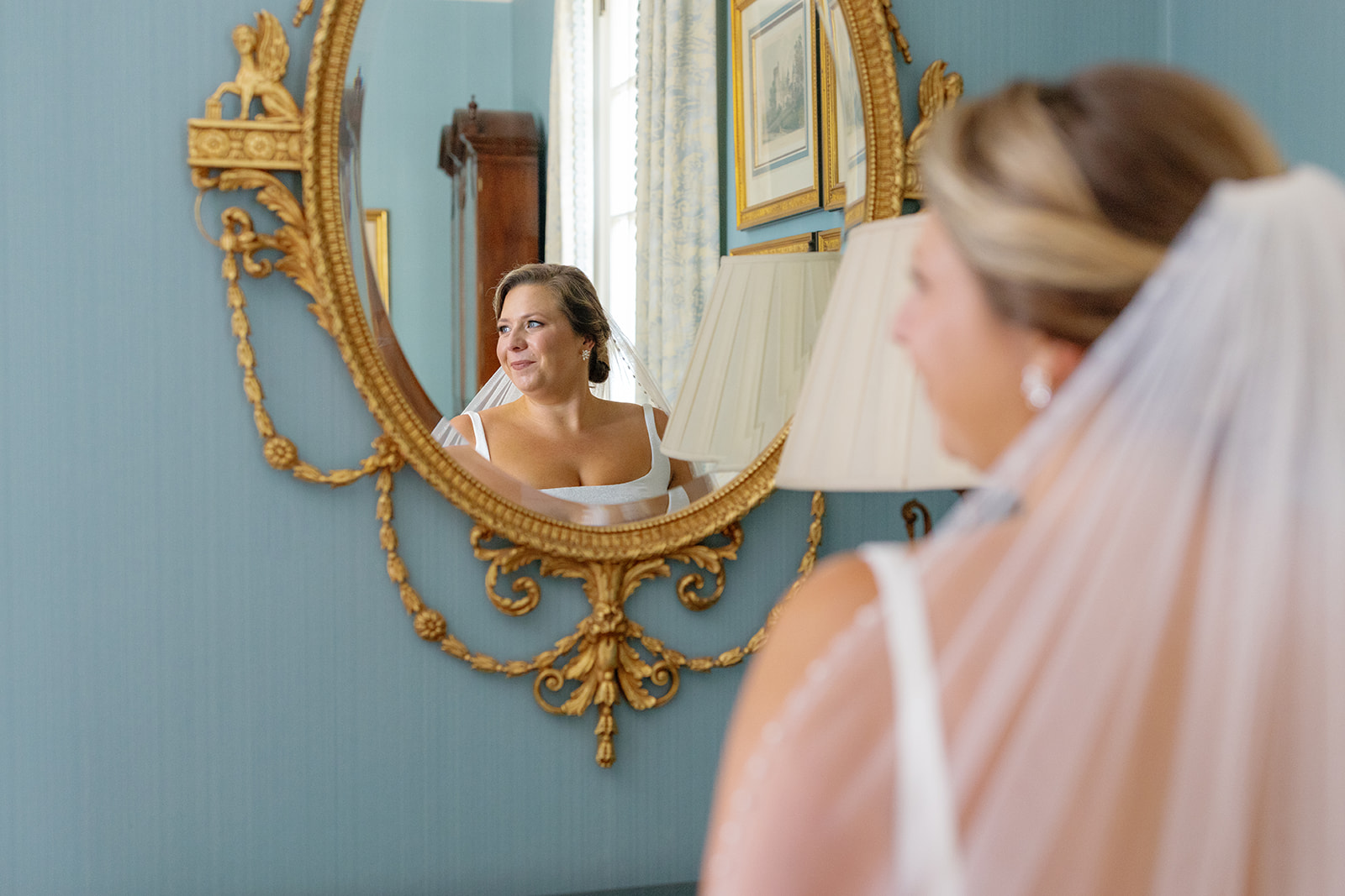 bride admires her reflection in an ornate gold mirror, glowing with bridal elegance.