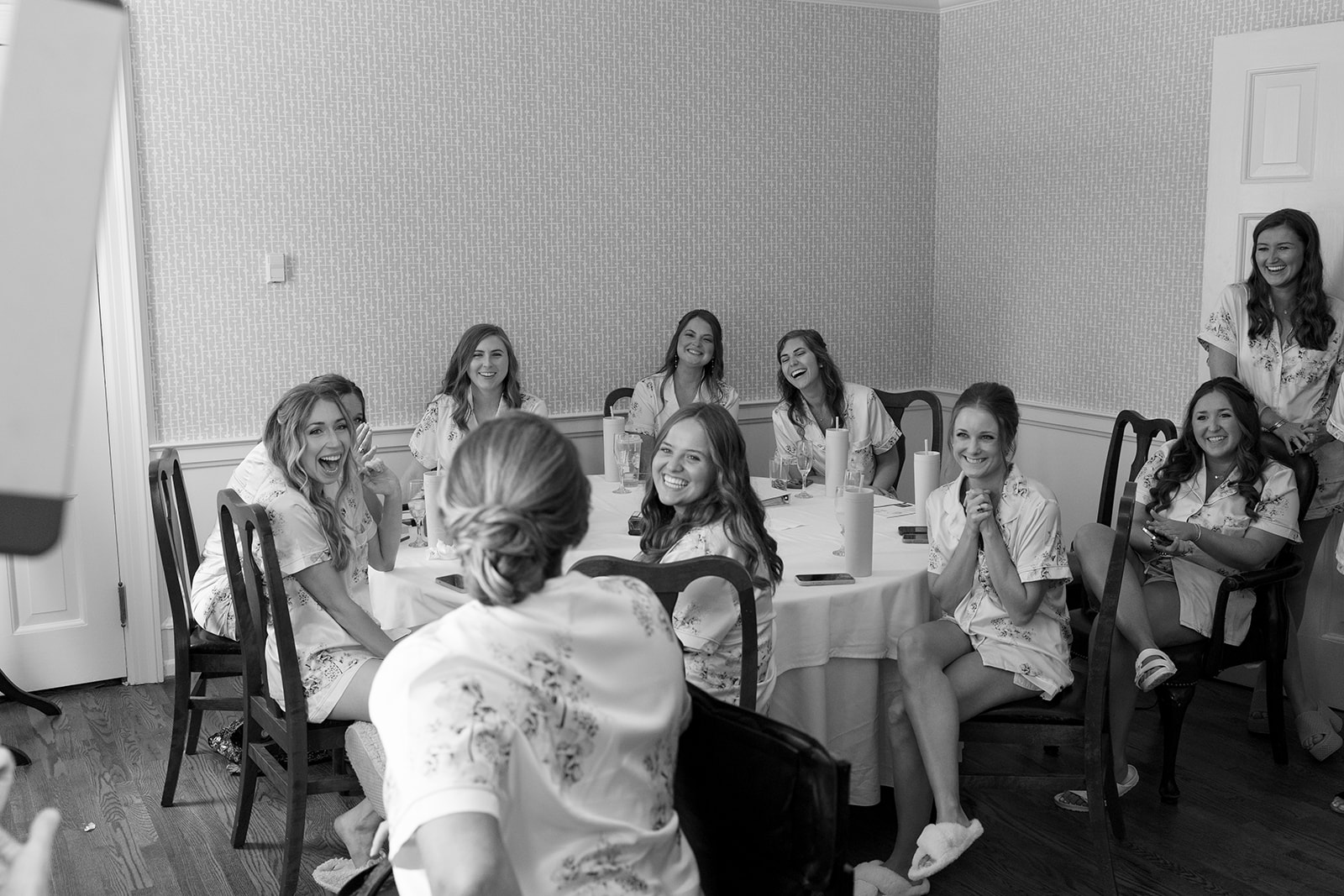 Bridesmaids burst into laughter as they sit around a table in matching floral pajamas.