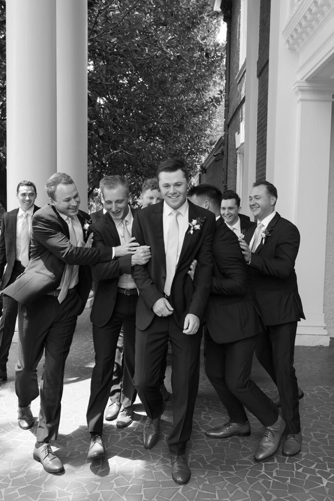 Groom shares a laugh with his groomsmen outside the venue in a playful, candid moment.