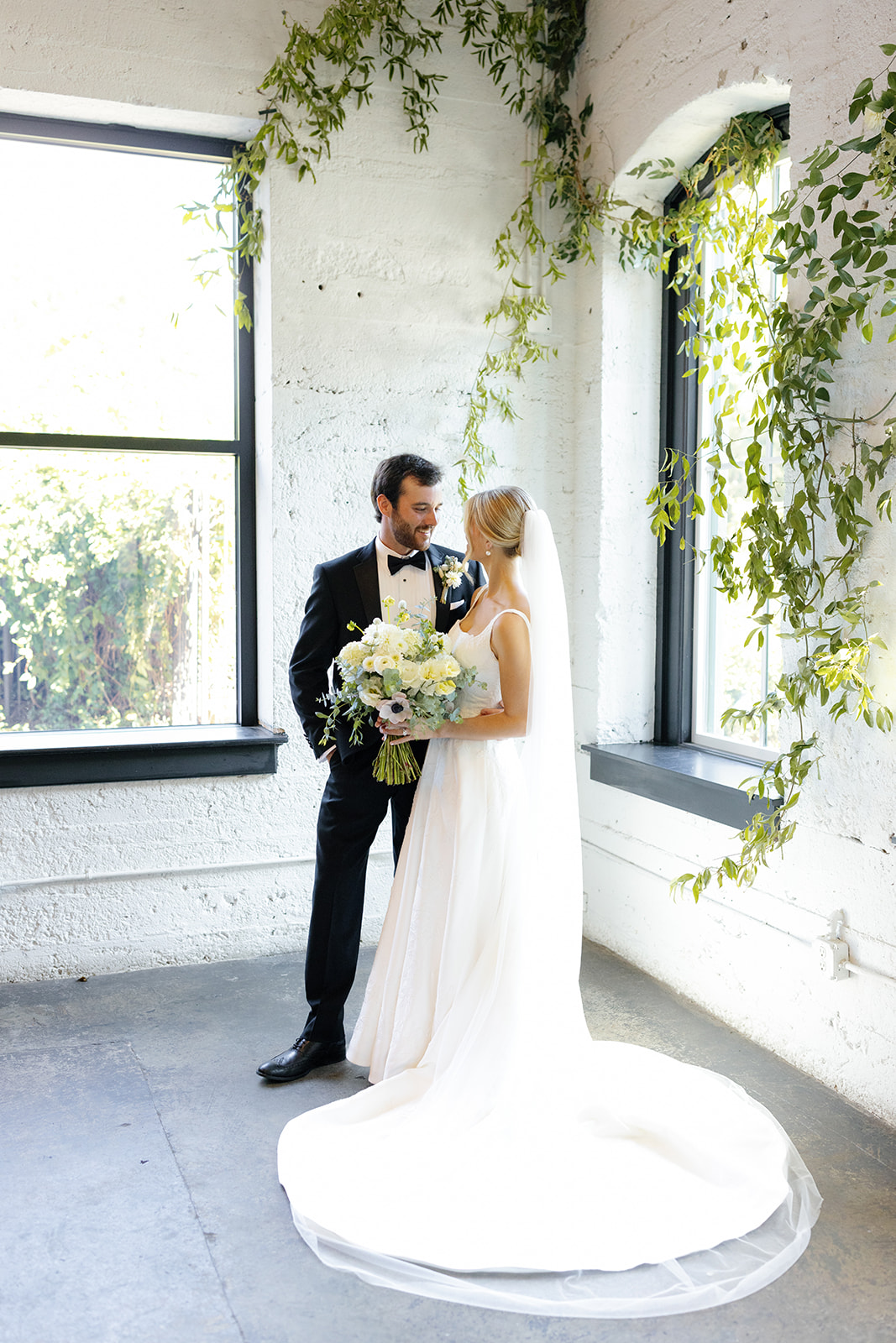 Bride and groom share a quiet, romantic moment by sunlit windows draped in greenery at their modern industrial venue.
