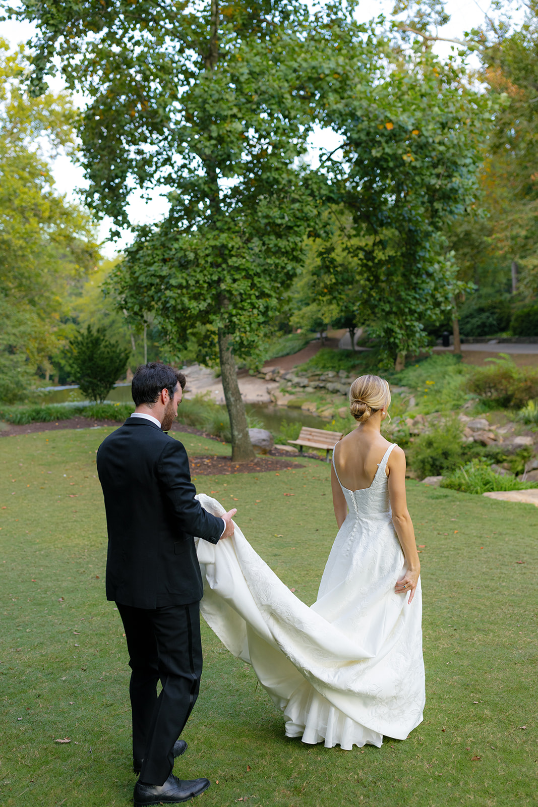Groom gently lifts the bride’s train as they stroll through lush greenery in Falls Park.