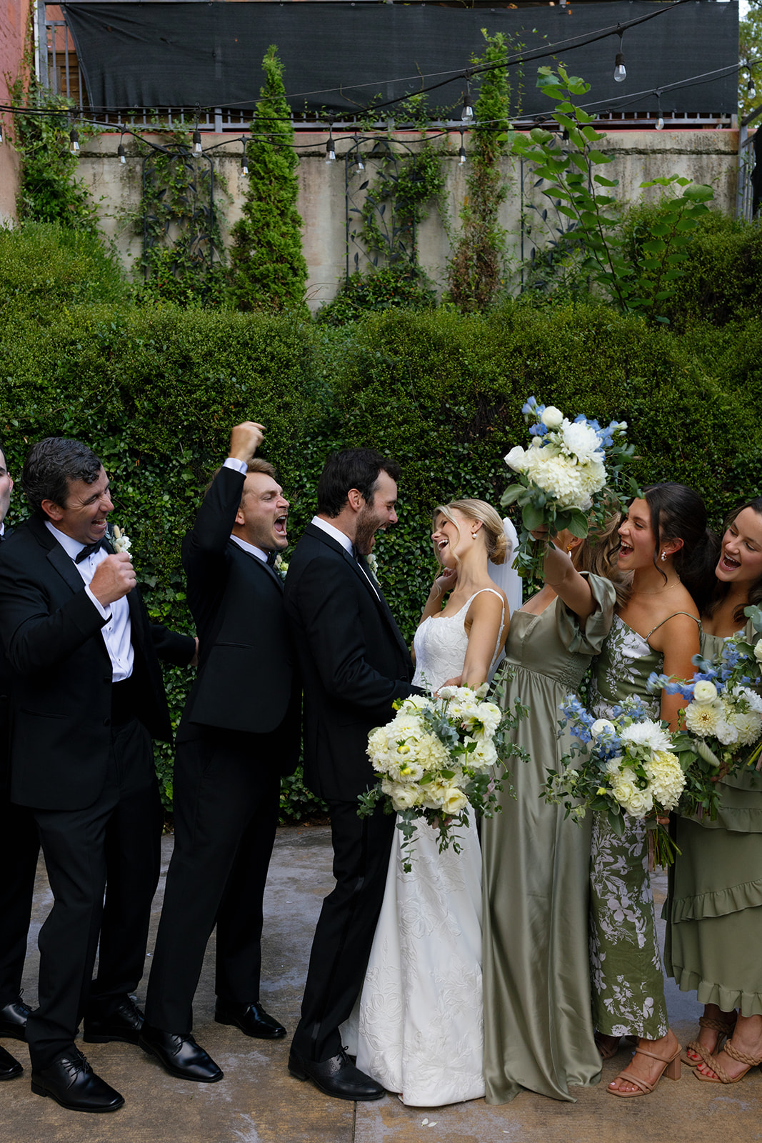 The couple celebrates surrounded by laughing friends, raising bouquets and fists in excitement.