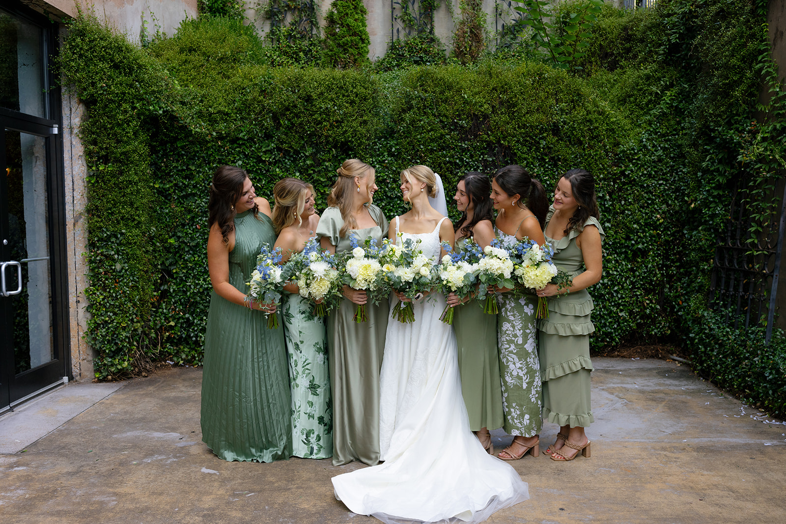 Bride smiles with her bridesmaids, all holding lush bouquets and wearing mix-and-match sage and green dresses.