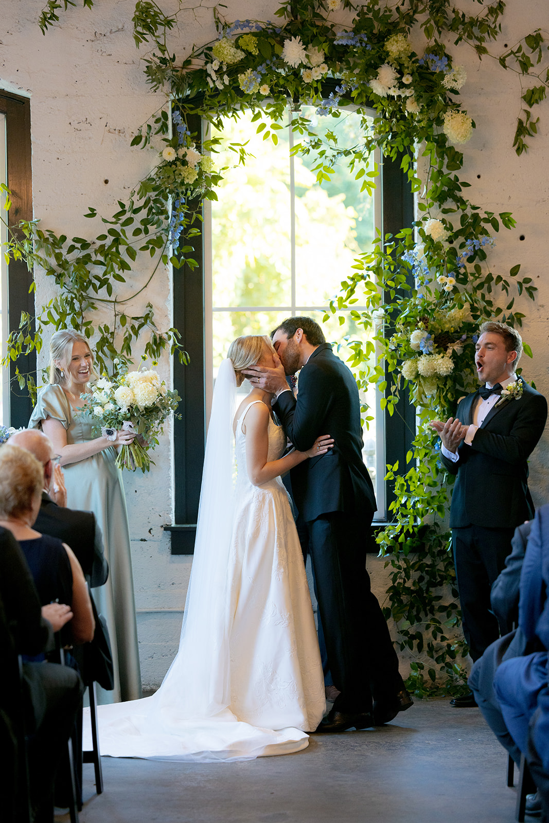 The couple shares their first kiss in front of a lush floral arch at their indoor Greenville wedding.