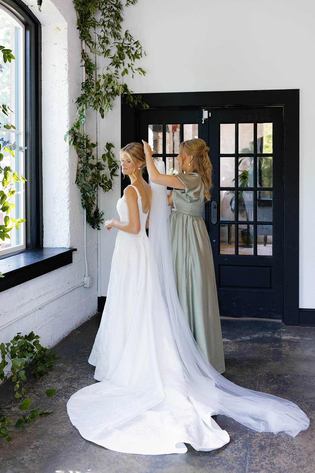 A bridesmaid gently places the bride’s veil in a light-filled room with lush greenery at Vue 1919.