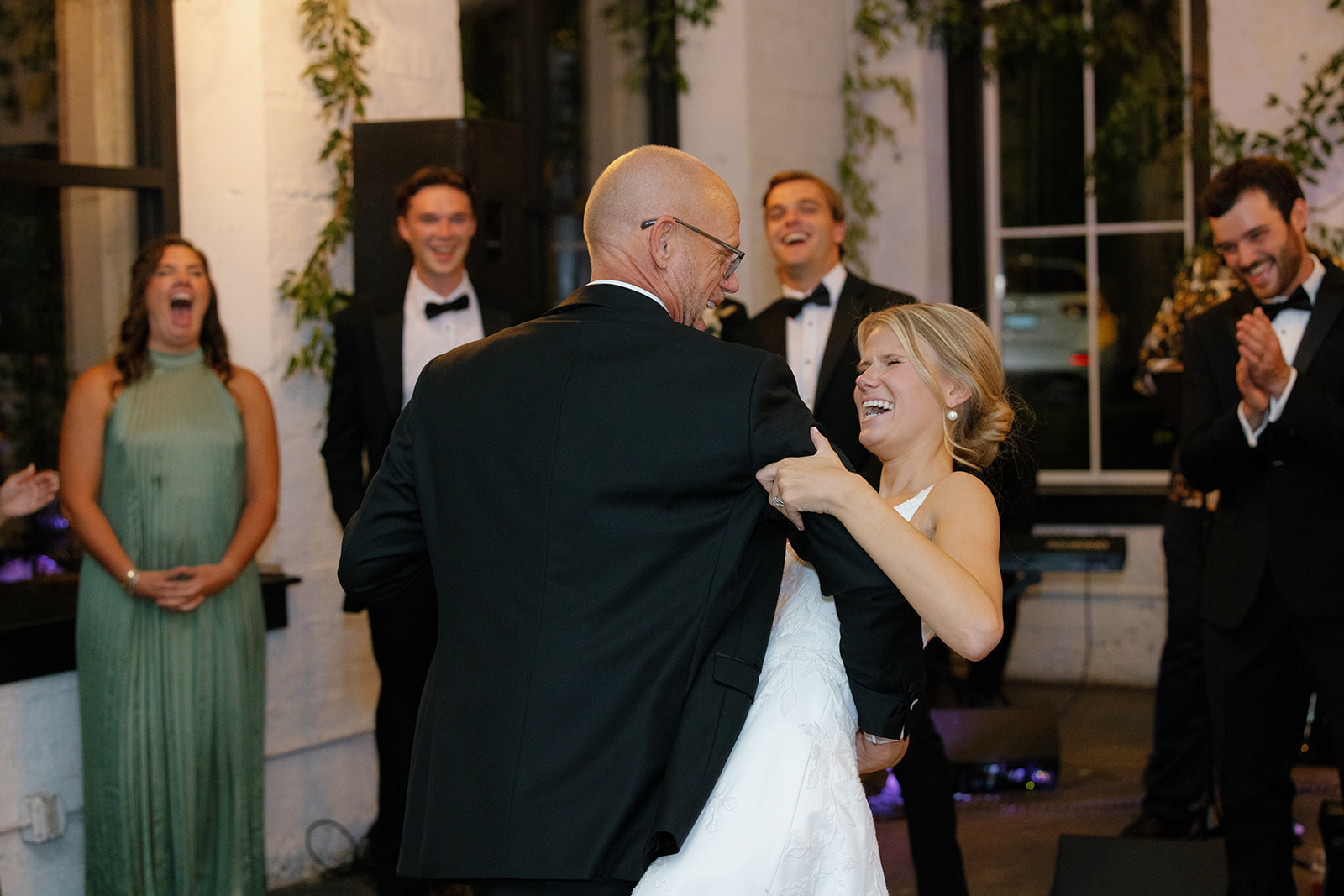 Bride shares a joyful dance with her father as wedding party looks on and laughs.