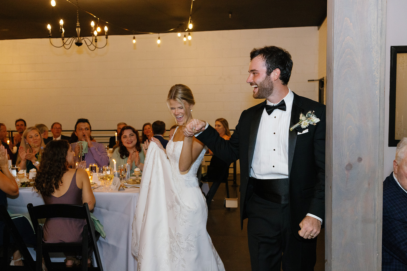 Bride and groom walk hand-in-hand through their reception, smiling as guests cheer them on.