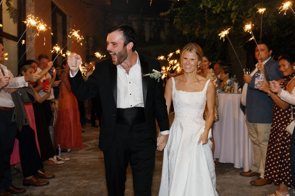 Excited groom high-fives a guest while leading his bride through a sparkler exit.