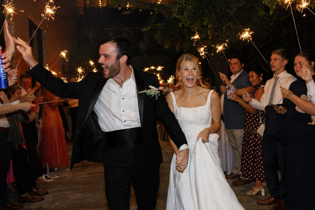 Groom pumps his fist as he and the bride walk through a sparkler tunnel of smiling guests.