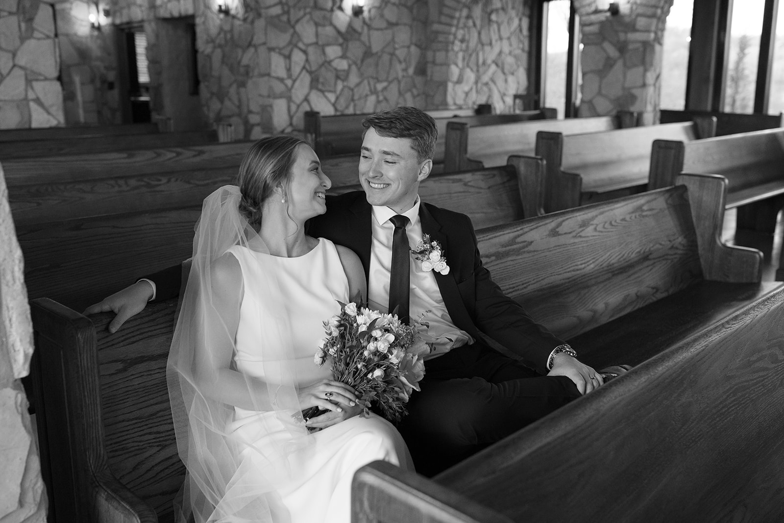 Newlyweds share a joyful laugh sitting side-by-side in a wooden church pew.
