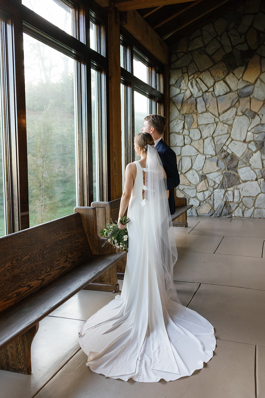 The couple looks out the large windows of glassy chapel, framed by stone walls and soft natural light.