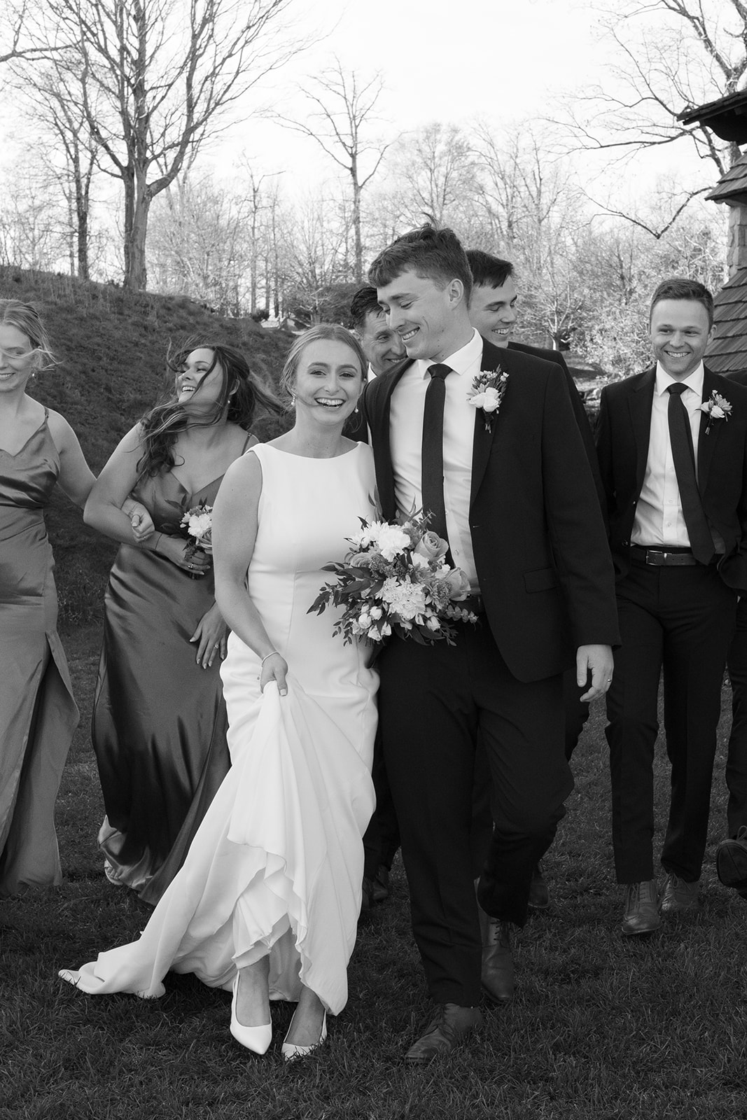 Bride grins toward the camera as the wedding party walks across the grass in celebration.