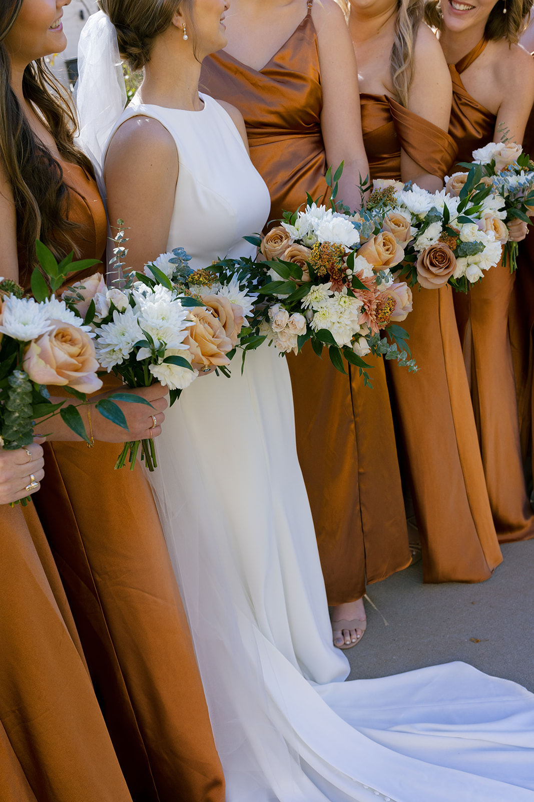 Bride and bridesmaids hold lush autumn-toned bouquets in satin gowns for a timeless bridal party portrait.