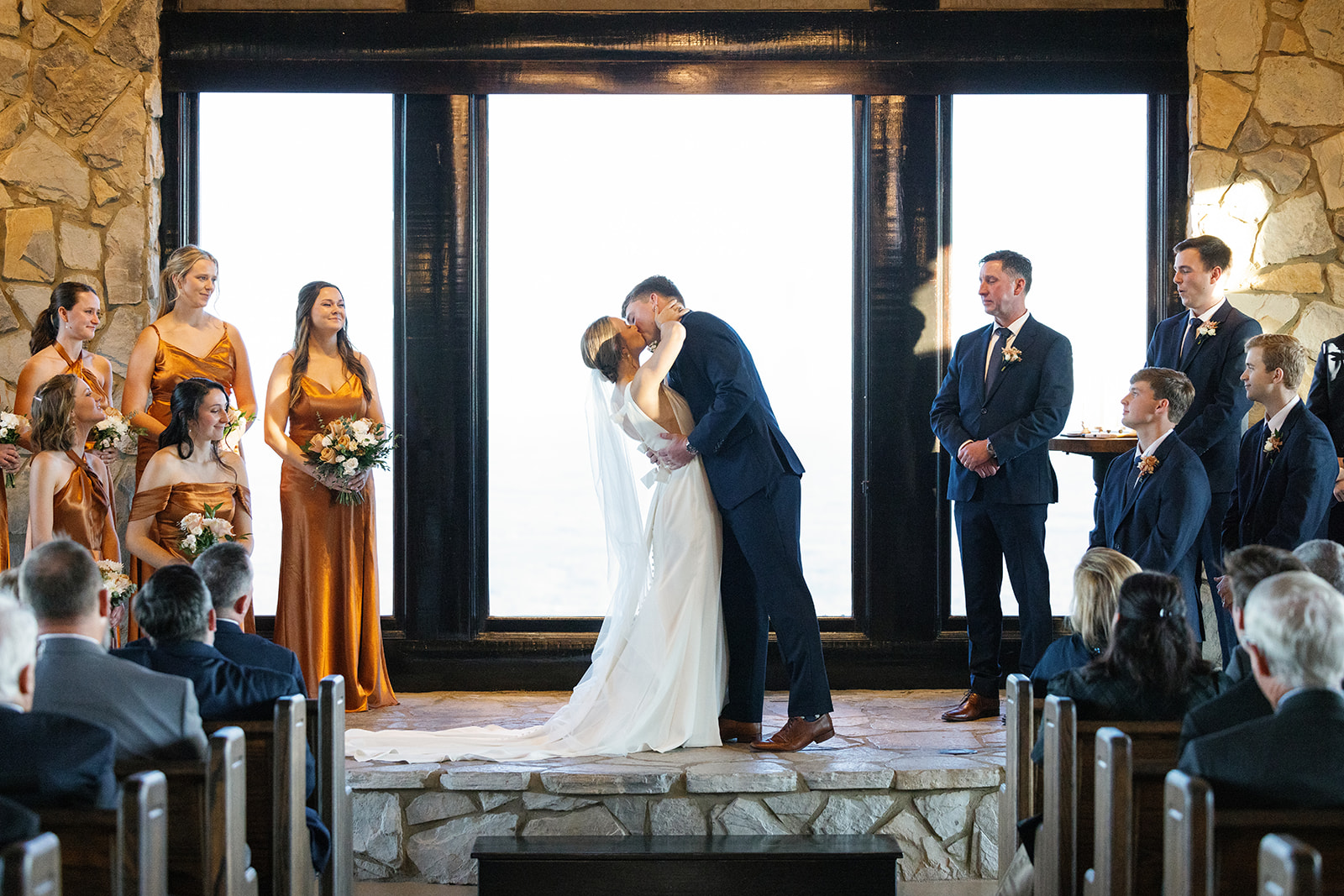 Bride and groom share their first kiss in front of floor-to-ceiling windows during their romantic indoor ceremony at glassy chapel.