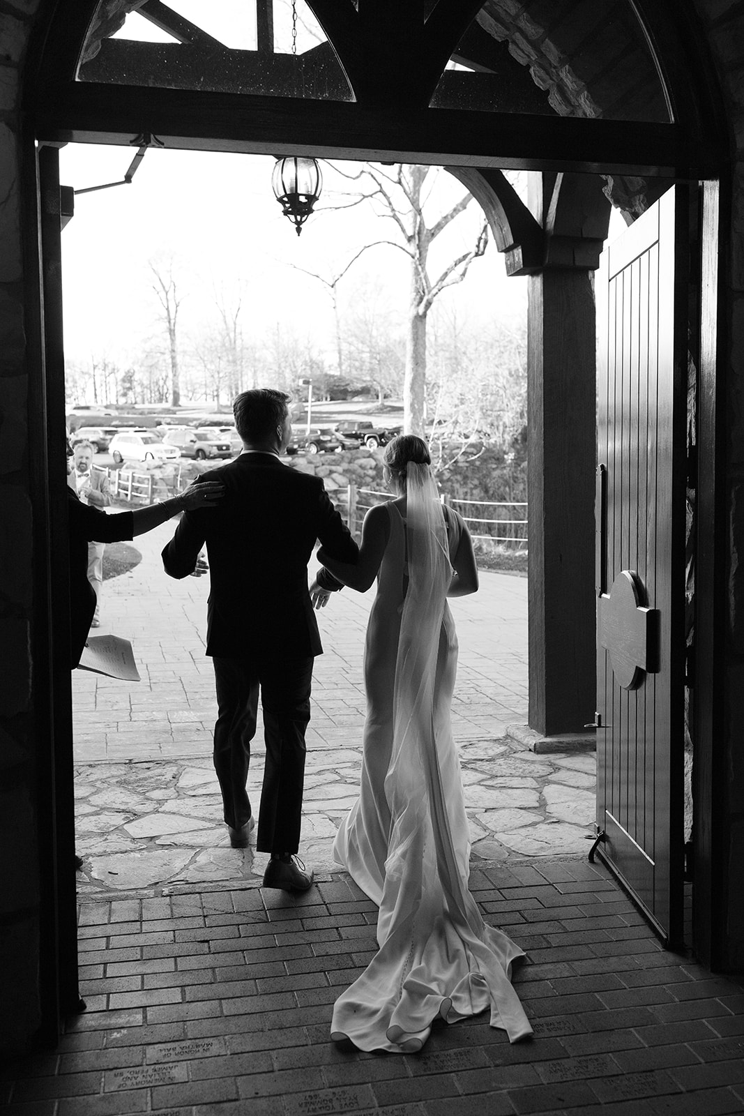 The couple walks hand-in-hand out of glassy chapel, framed by rustic open doors and soft afternoon light.