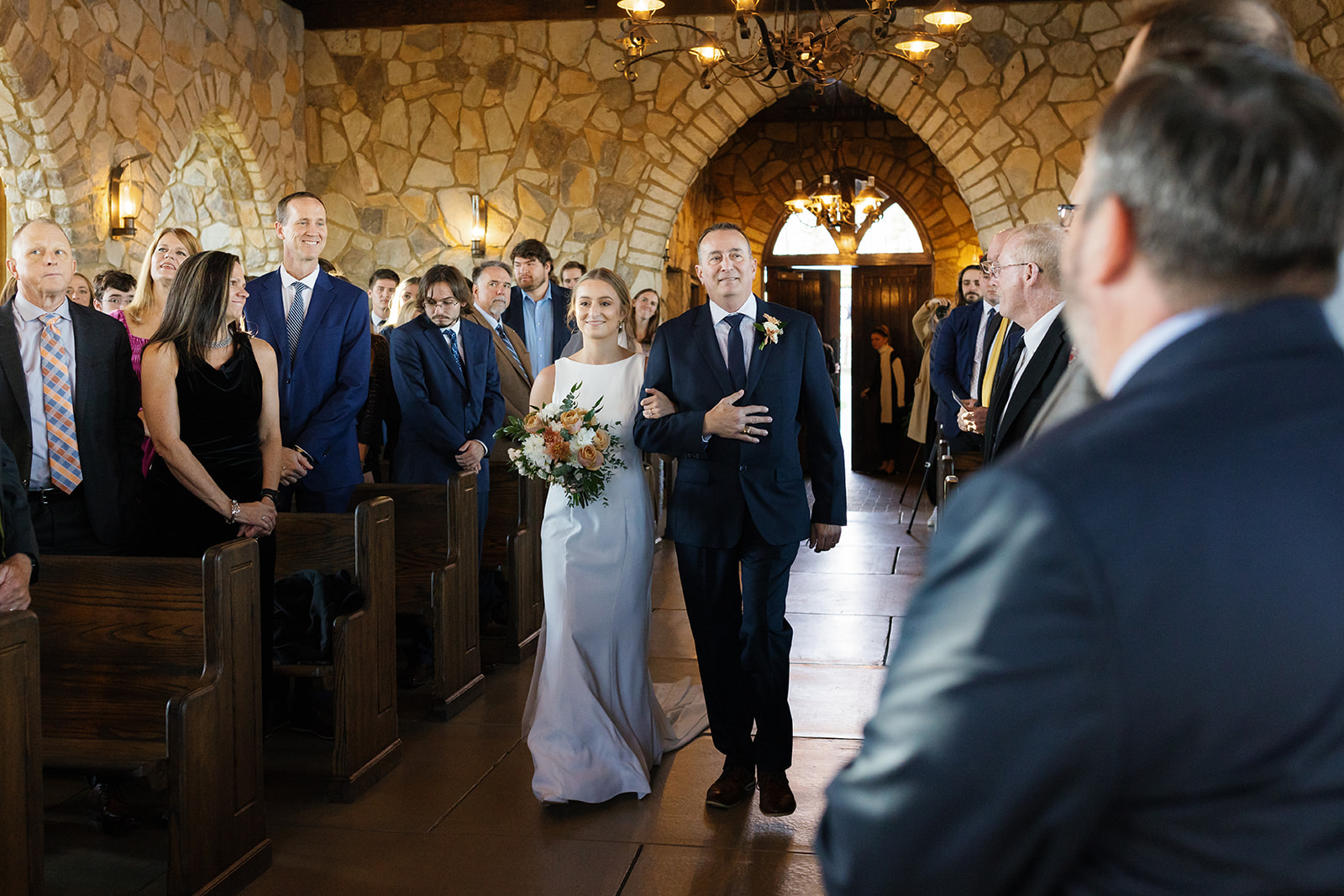 Bride walks arm-in-arm with her father toward the groom in a romantic moment inside glassy chapel.