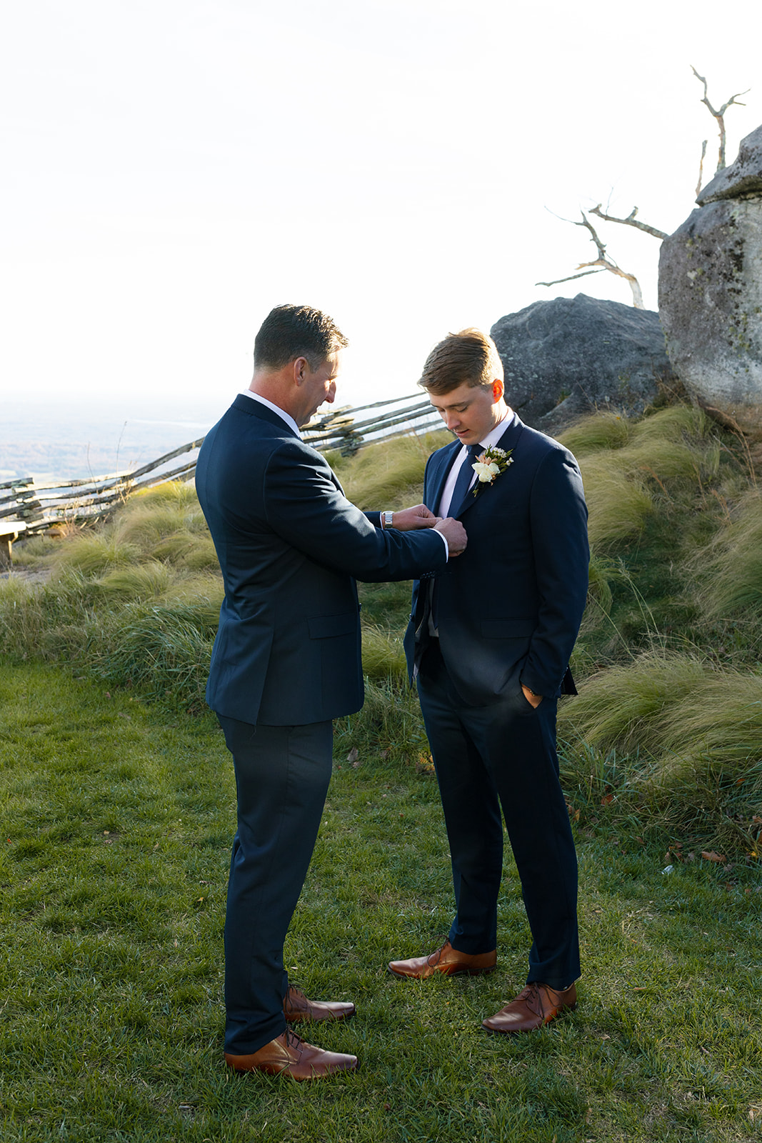 The father of the groom helps fix the groom’s boutonnière on a grassy overlook before the ceremony.
