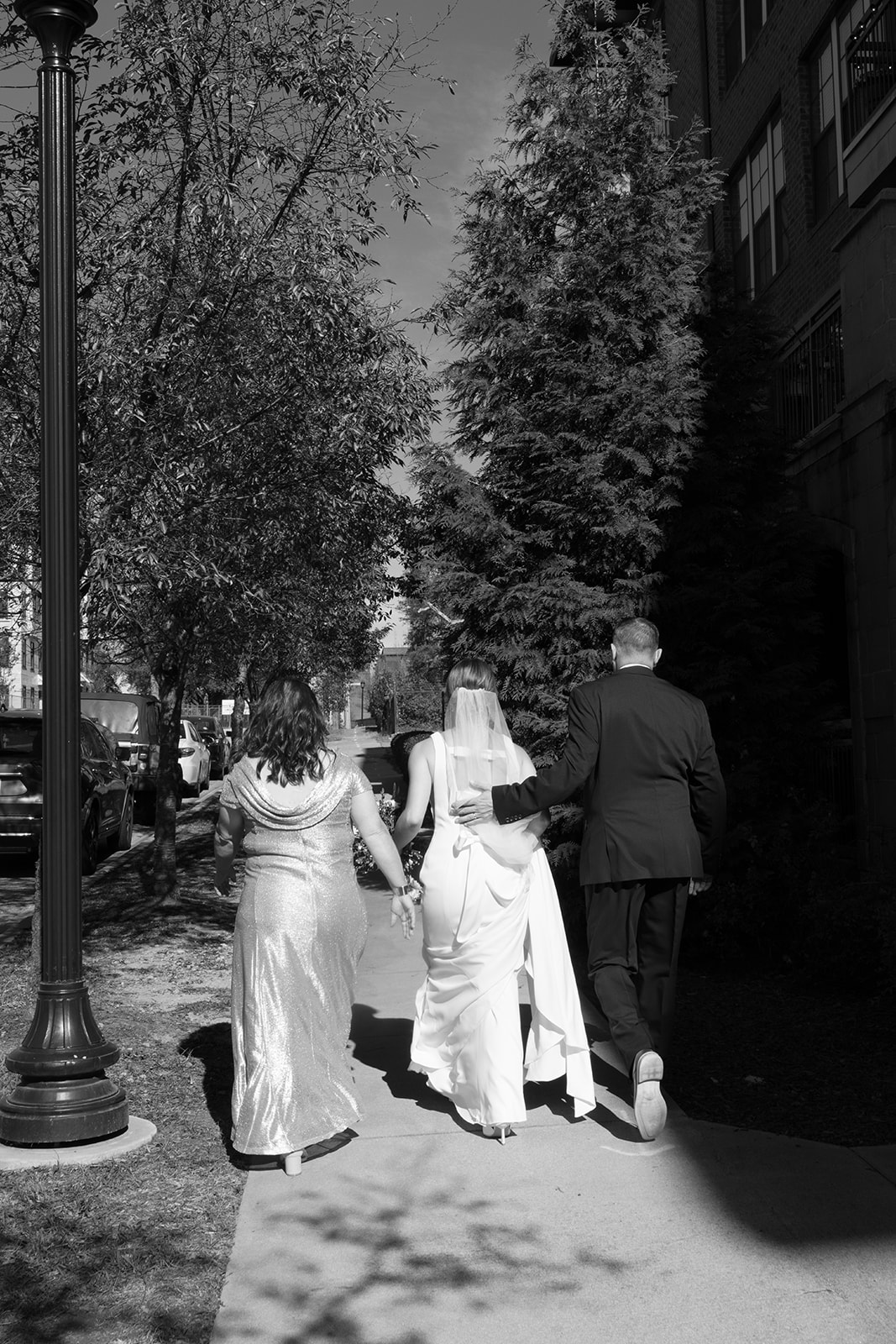 Bride walks arm-in-arm with her parents down a sun-dappled sidewalk toward the ceremony.