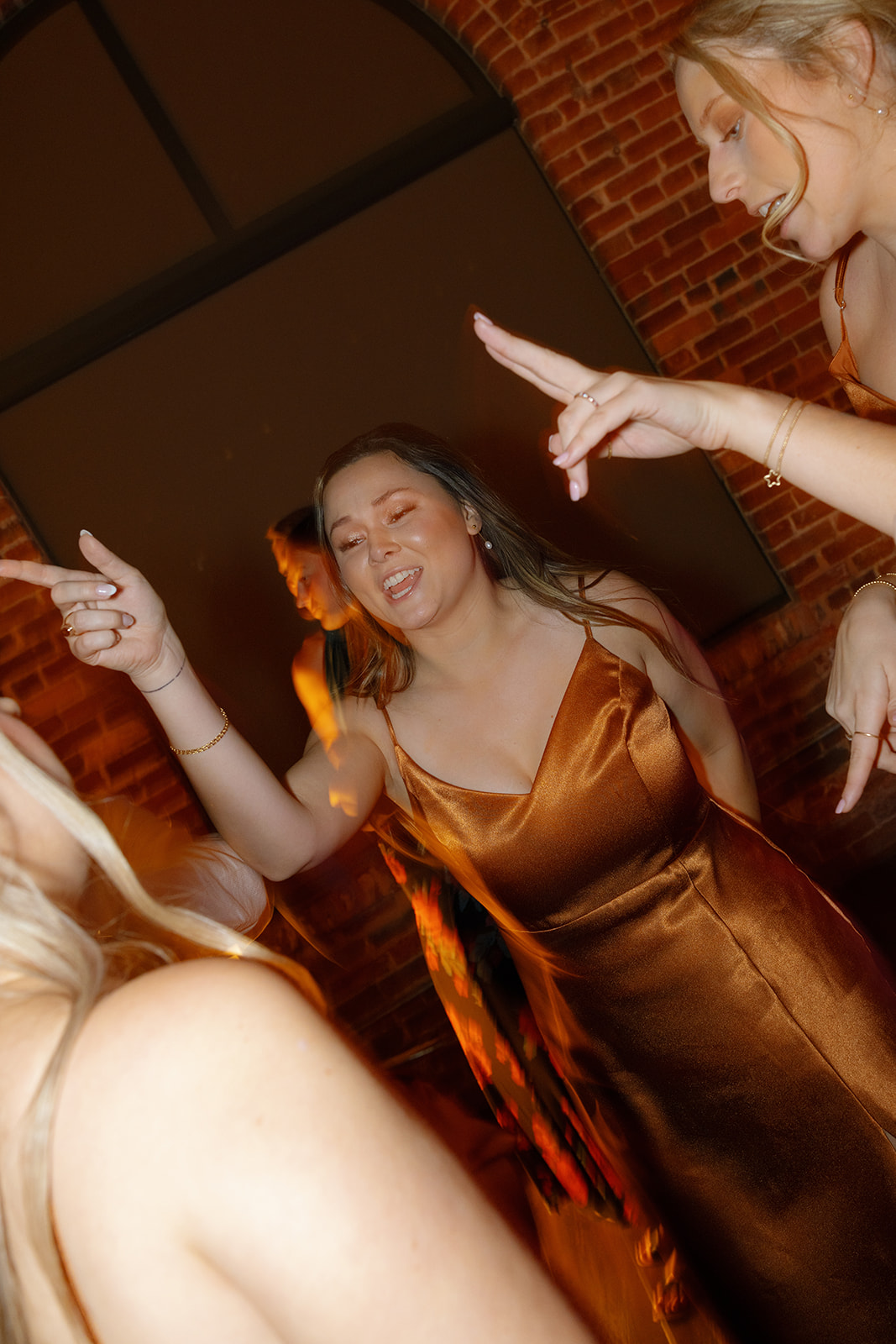 Bridesmaid in a copper satin dress dances with joy against the brick walls of the reception venue.